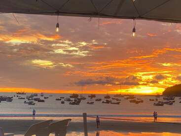 Um pôr do sol vibrante pinta o céu de laranja e amarelo sobre uma baía calma repleta de barcos ancorados, vistos de baixo de um dossel externo com luzes penduradas.