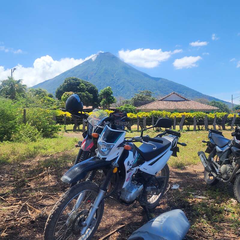 Três motocicletas estão estacionadas em uma área gramada, com um cenário de um vulcão, vegetação exuberante, uma casa com telhado de telha e céu azul.