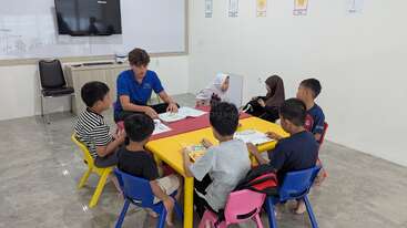 Un enseignant est assis à une table jaune avec sept enfants et les fait participer à une activité de lecture. La salle de classe est lumineuse et des affiches éducatives sont accrochées au mur.