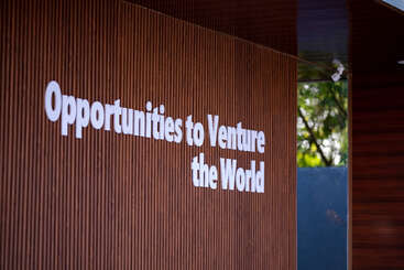 Cette image montre un mur en bois sur lequel on peut lire en gras et en blanc "Opportunities to Venture the World". Des arbres et des paysages extérieurs sont visibles à travers une fenêtre.
