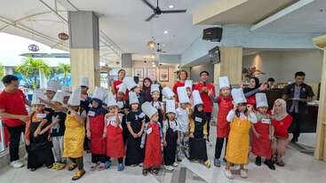 Un groupe d'enfants portant des tabliers et des toques pose ensemble à l'intérieur avec plusieurs adultes. Le cadre semble festif, probablement celui d'un cours de cuisine ou d'un atelier.