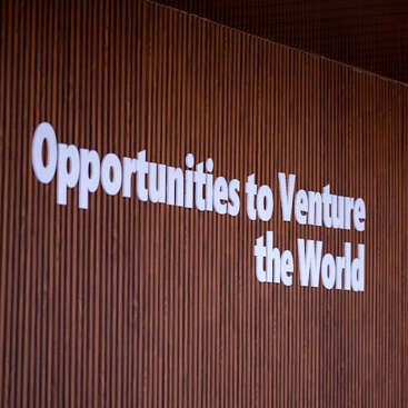 This image shows a wooden wall featuring bold, white text that reads, "Opportunities to Venture the World." Trees and outdoor scenery are visible through a window.