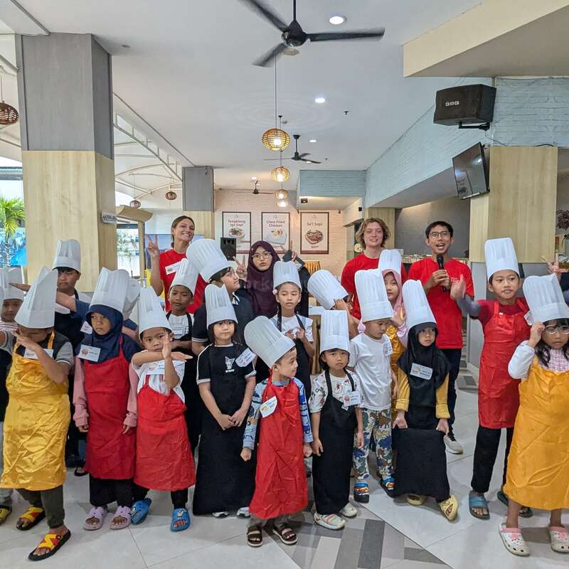 A group of children wearing aprons and chef hats pose together indoors with several adults. The setting appears festive, likely a cooking class or workshop event.