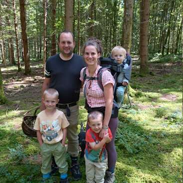Uma família de cinco pessoas posa em uma floresta exuberante. A mãe carrega uma criança pequena em uma mochila. Todos sorriem, desfrutando juntos de uma aventura na natureza ao ar livre.