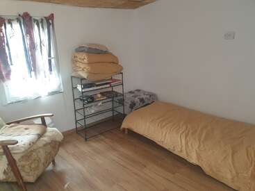 A simple room with a single bed, mustard bedding, armchair, metal shelf with folded blankets, books, a small table, wooden floor, and a window with curtains.