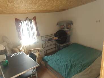 This cozy, small room includes a bed with a green cover, a desk with a chair, armchair, window with curtains, shelving unit, and stacked storage bins.