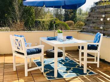 Um pátio ao ar livre cheio de estilo tem uma mesa branca, cadeiras com almofadas azuis, um guarda-sol azul, um arranjo floral no centro da mesa, um tapete estampado e um cenário natural — perfeito para uma refeição relaxante sob o sol.