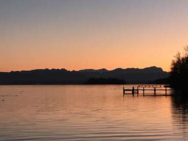 Um lago calmo ao pôr do sol, tons tranquilos de laranja e rosa no céu, montanhas e um cais recortados contra o horizonte, águas tranquilas refletindo a luz, árvores emoldurando a cena serena.