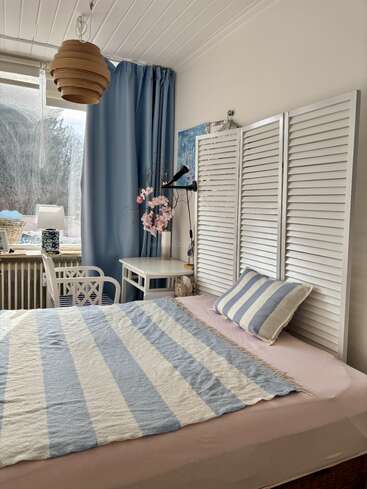 Cette chambre chaleureuse est décorée dans des tons bleu pastel et blanc ; elle est équipée d'une parure de lit rayée, d'une suspension en bois, d'un bureau, de rideaux bleus, d'une fenêtre et de fleurs décoratives.