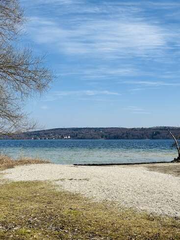 Une scène paisible au bord d'un lac, avec une eau bleue et calme, une rive caillouteuse, de l'herbe clairsemée, des branches d'arbres dénudées, des collines au loin, des maisons éparpillées et un ciel partiellement nuageux.