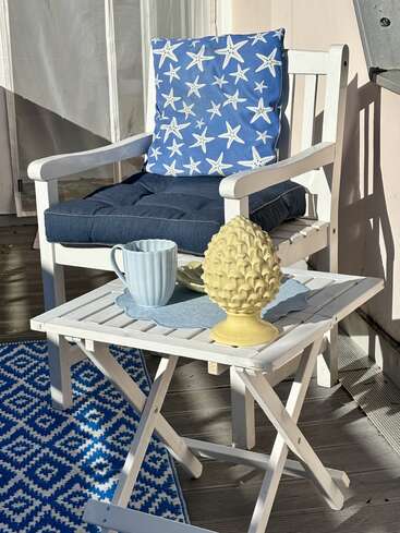 Une scène chaleureuse sur une terrasse, avec une chaise blanche, un coussin à motifs d'étoiles de mer, une petite table, une tasse bleue, un objet décoratif jaune et un tapis à motifs bleus, le tout baigné de soleil.