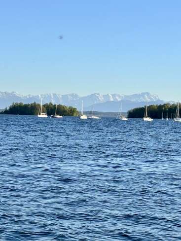 Um lago azul cristalino com ondas suaves, vários veleiros flutuando, ilhas verdes e majestosas montanhas cobertas de neve ao fundo, sob um céu claro e sem nuvens.