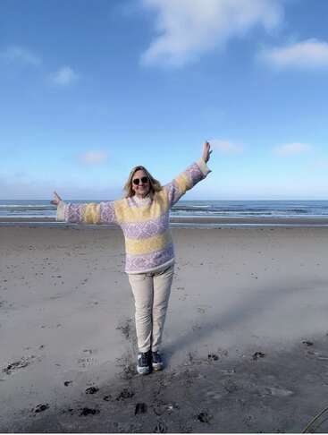 Uma mulher está alegremente em pé numa praia de areia, com os braços abertos, usando óculos escuros e um suéter colorido. O céu está limpo e azul acima dela.