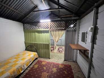 Um quarto rústico simples com uma cama de solteiro, paredes de madeira, teto de lata, tapetes estampados, uma pequena prateleira, uma porta verde e uma cortina que leva a outro cômodo.