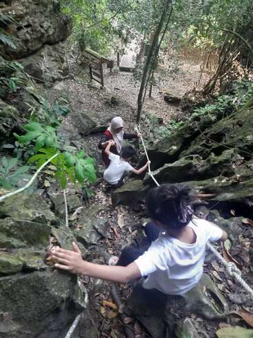 Three people, including a woman in a headscarf and two children, carefully climb down a steep, rocky hillside using ropes, surrounded by dense, green forest.
