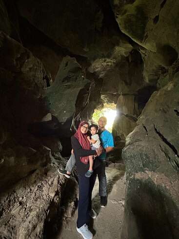 A family of three stands together inside a rocky cave, smiling at the camera. Sunlight streams through the entrance, creating a warm and adventurous atmosphere.