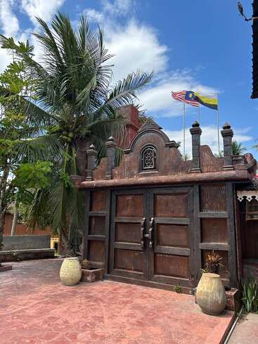A traditional wooden gate stands tall, flanked by lush tropical trees and large ceramic pots. Above, Malaysian flags wave against a beautiful, clear blue sky.