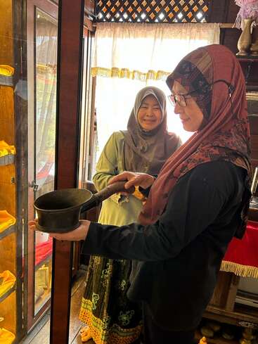 Two women in traditional attire stand indoors. One woman holds an antique metal pot, examining it closely, while the other woman smiles warmly in the background.