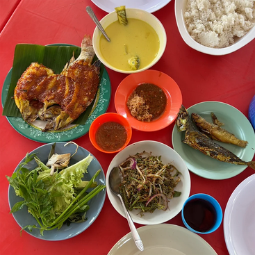 A colorful meal spread featuring grilled fish, fried fish, rice, vegetables, yellow curry, salad, sambal, and a drink, all placed on a red table.