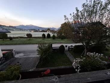 Eine friedliche, frühmorgendliche Szene mit einem malerischen Garten, einer ruhigen Straße, frostigem Gras, fernen Häusern und majestätischen Bergen unter einem weichen, blauen Himmel mit Pastellfarben.
