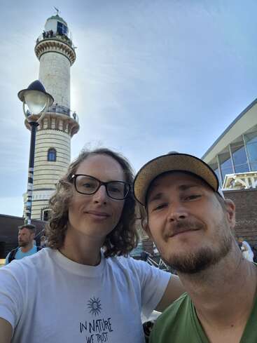 Ein lächelndes Paar macht ein Selfie im Freien mit einem hohen Leuchtturm im Hintergrund. Das Wetter ist sonnig, und andere Menschen sind in der Nähe zu sehen.