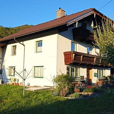 Dieses Bild zeigt ein charmantes Haus auf dem Land mit einem roten Ziegeldach, einem Holzbalkon und grün gestrichenen Fenstern, umgeben von Gras, Blumen und in der Sonne liegend.