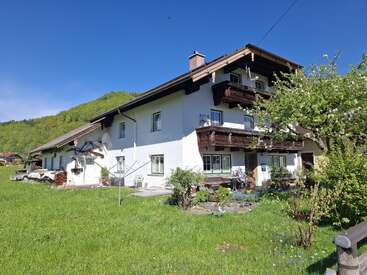 Ein charmantes, großes weißes Bauernhaus mit Holzbalkonen steht inmitten von saftig grünem Gras und Bäumen unter einem strahlend blauen Himmel, umgeben von malerischen sanften Hügeln.