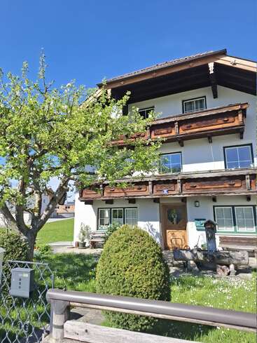 Ein charmantes, traditionelles Haus mit Holzbalkonen steht unter einem strahlend blauen Himmel. Ein üppig belaubter Baum und gepflegte Sträucher schmücken den grünen Vorgarten. Eine friedliche, sonnige Szenerie.