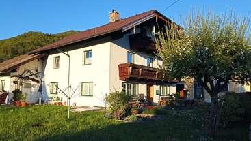 Dieses Bild zeigt ein charmantes Haus auf dem Land mit einem roten Ziegeldach, einem Holzbalkon und grün gestrichenen Fenstern, umgeben von Gras, Blumen und in der Sonne liegend.