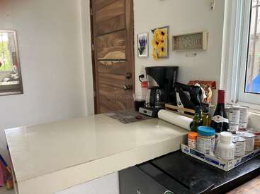 Un coin cuisine confortable avec une porte en bois, une cafetière, un assortiment d'articles de garde-manger, des bouteilles de vin, des conserves, des serviettes en papier et des décorations murales gaies près d'une fenêtre.