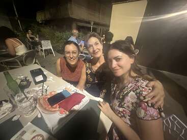Trois femmes sont assises près les unes des autres, souriantes, à une table en plein air, la nuit. De la nourriture, des boissons et un smartphone sont posés sur la table, suggérant une réunion festive.