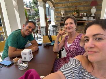Trois personnes sont assises à la table d'un café en plein air et sourient chaleureusement à l'appareil photo. La lumière du soleil éclaire leurs visages. Des boissons, des téléphones et du sucre sont posés sur la table. Ambiance détendue.