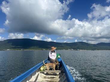 A person wearing a hat sits in a small blue boat with a dog, moving across calm lake waters, mountains and dramatic clouds in the background.