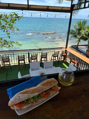 A fresh sandwich and glass of white wine sit on a rustic table, overlooking a scenic beachside patio with ocean views, palm trees, and clear skies.
