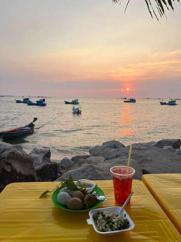 A peaceful seaside scene at sunset, colorful fishing boats on calm water, food and drink on a yellow table, rocks nearby, palm leaves overhead, tranquil atmosphere.
