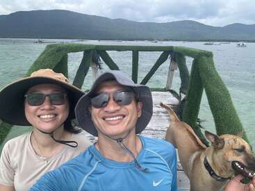 A man and woman in sun hats and sunglasses smile on a wooden pier, accompanied by a happy dog. The background features water and green mountains.