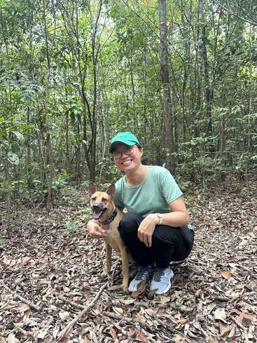A woman wearing a green cap and glasses kneels beside a happy brown dog in a lush forest, surrounded by trees and dry leaves on the ground.