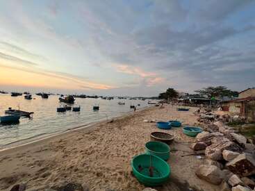 Several round fishing boats rest on a sandy beach, with rocky edges and calm sea waters. Countless boats float offshore under a dramatic, colorful sky at sunset.