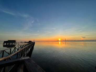 A wooden pier extends over calm water at sunrise, with tranquil reflections, clear blue sky, soft clouds, and two huts on the serene horizon. Peaceful ambiance.