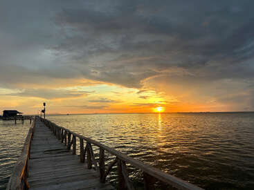 A wooden pier stretches into the calm sea under a dramatic, colorful sunset sky. The reflection of the sun glows on rippling water, creating tranquility.
