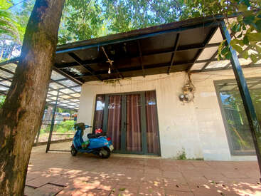 A blue scooter and helmet are parked outside a small house with glass doors. The building features a black metal roof, surrounded by trees and greenery.
