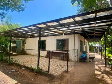 A small, simple house with a large metal awning, fenced patio, blue scooter, two black dogs, and surrounded by lush green trees under a sunny sky.