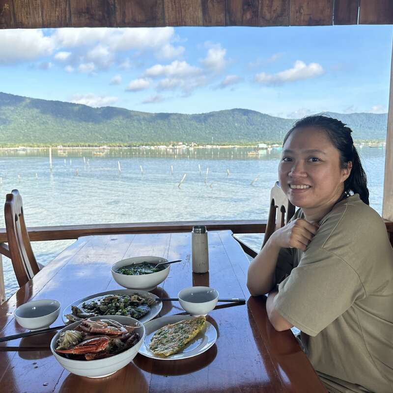 A woman smiles at a wooden table set with seafood and soup, enjoying a meal outdoors with a scenic backdrop of mountains, water, and blue sky.