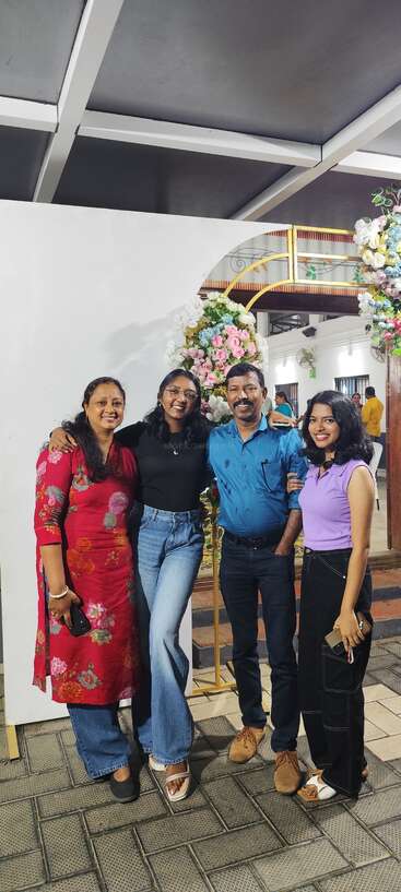 Four people are smiling, posing together in front of a floral arch backdrop. They look happy and festive, likely at a celebration or special event indoors.