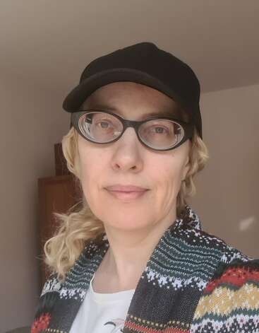 A woman with blonde hair wears a black cap, glasses, and a patterned sweater. She stands indoors with neutral walls, softly lit by natural sunlight.