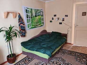 A cozy bedroom features a green bed, wall tapestry of a forest, potted plant, patterned rug, hanging coat, small shelf, and photos on cream walls.