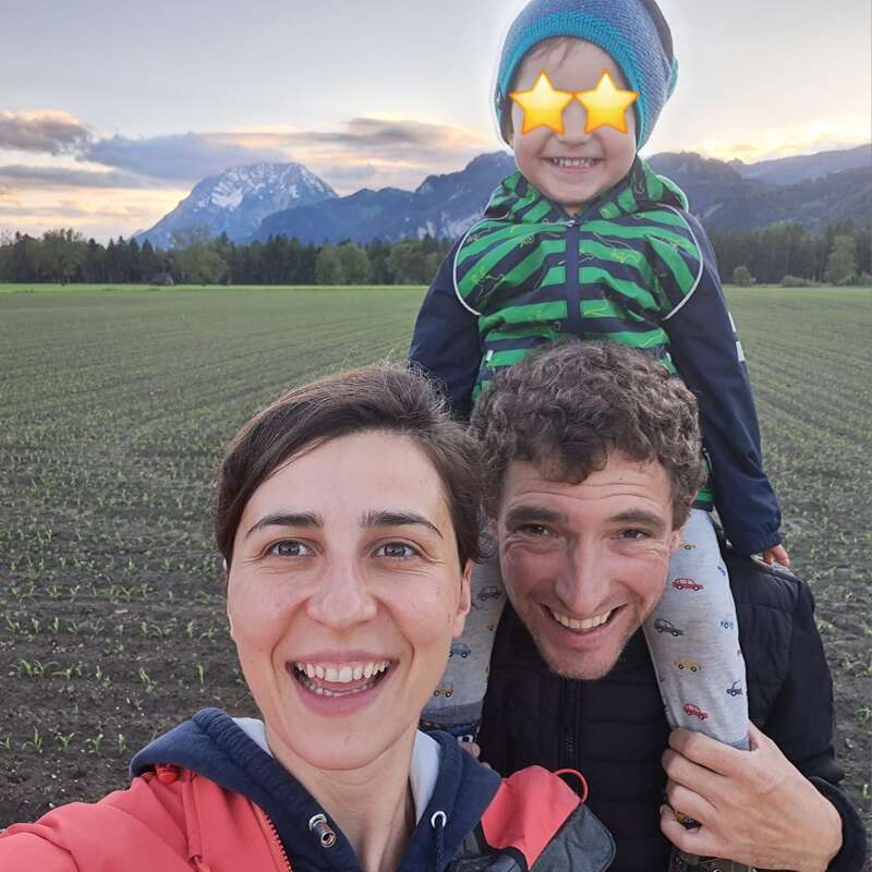 A happy family takes a cheerful selfie outdoors in a field, with mountains and a sunset in the background. The child’s eyes are covered with star emojis.