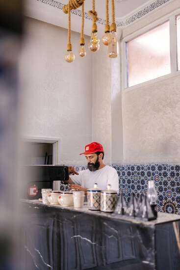 A bearded man in a red cap makes coffee behind a counter with ceramic cups in a stylish cafe featuring patterned tiles and decorative hanging light bulbs.