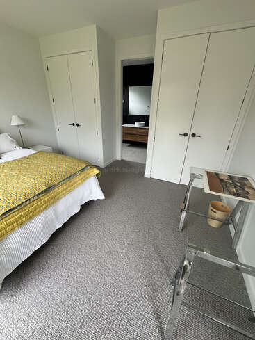 Un dormitorio moderno y minimalista con moqueta gris claro, paredes blancas, armarios dobles, un escritorio de cristal y una colcha amarilla. Cuarto de baño visible a través de la puerta.