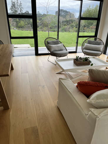 Una acogedora sala de estar con suelo de madera clara, sillas modernas, un sofá blanco con cojines naranjas y grandes ventanales que ofrecen una vista panorámica de un verde jardín.